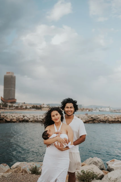 A family portrait with the city behind them | Barcelona newborn family photoshoot The mother holding the newborn with the father close behind and the city across the water during their newborn family photoshoot in Barcelona.
