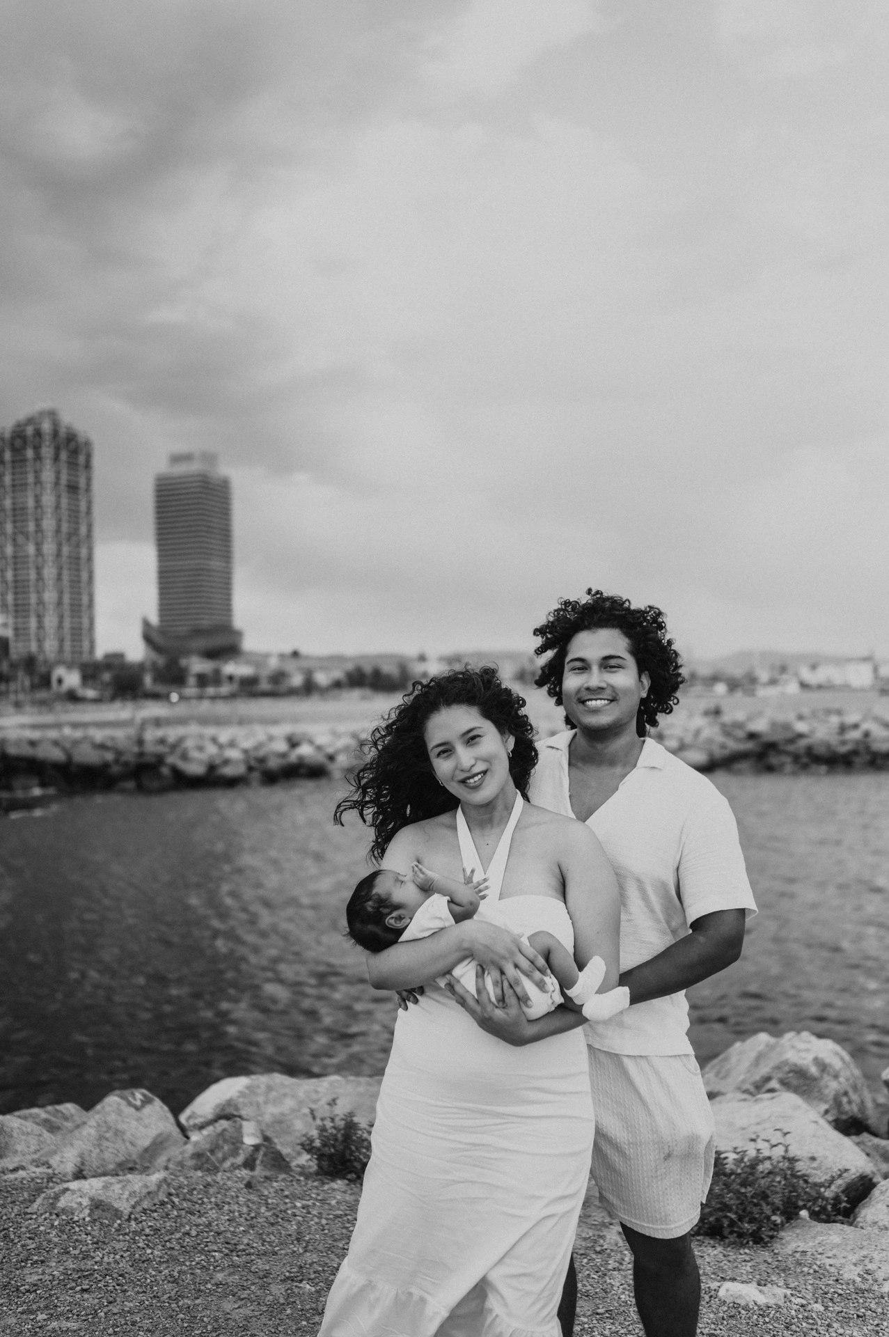 Black-and-white photograph: the mother holding the newborn with the father close behind and the city across the water during their newborn family photoshoot in Barcelona.