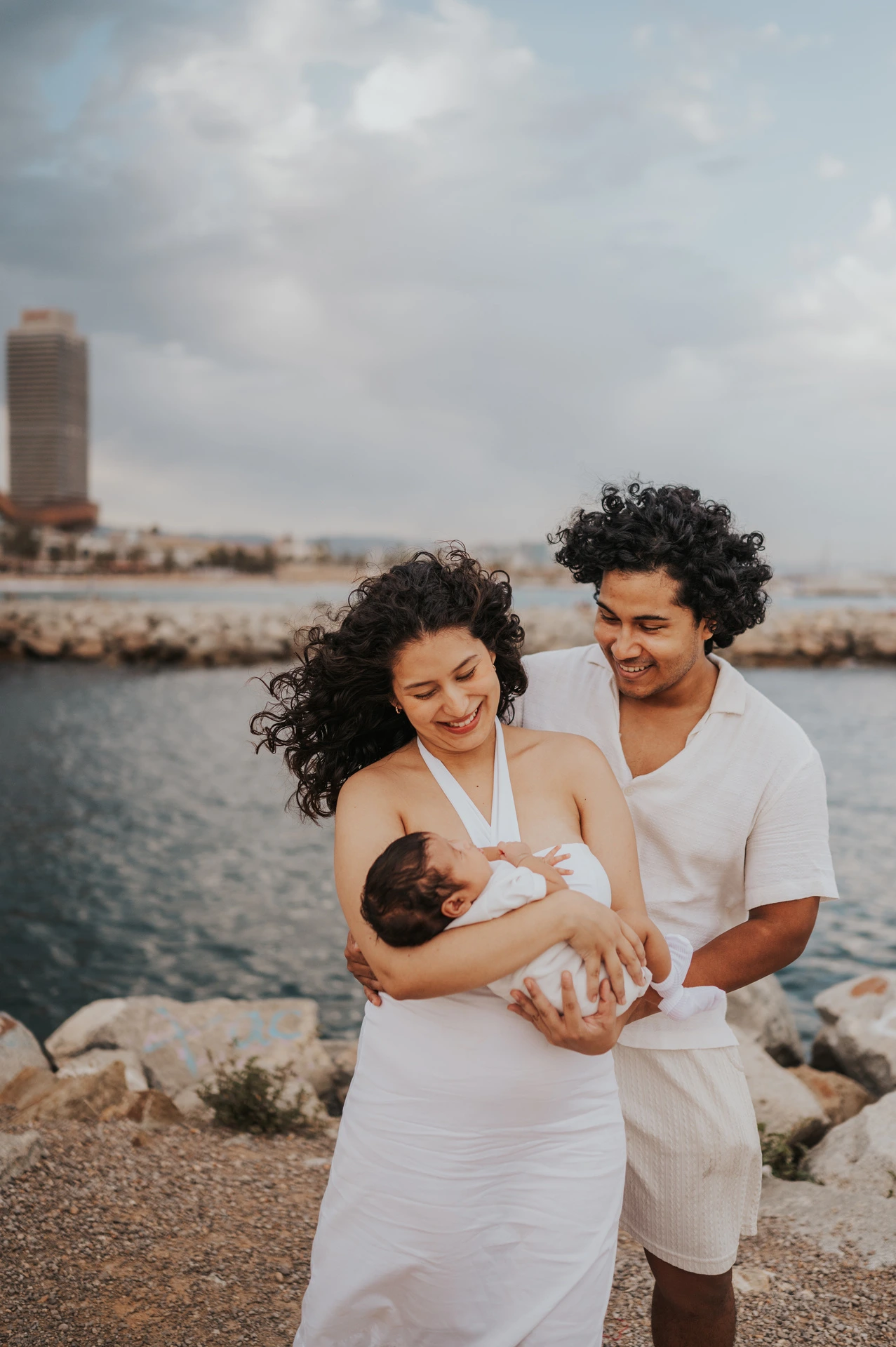 The mother holding the newborn with the father close behind and the city across the water during their newborn family photoshoot in Barcelona.