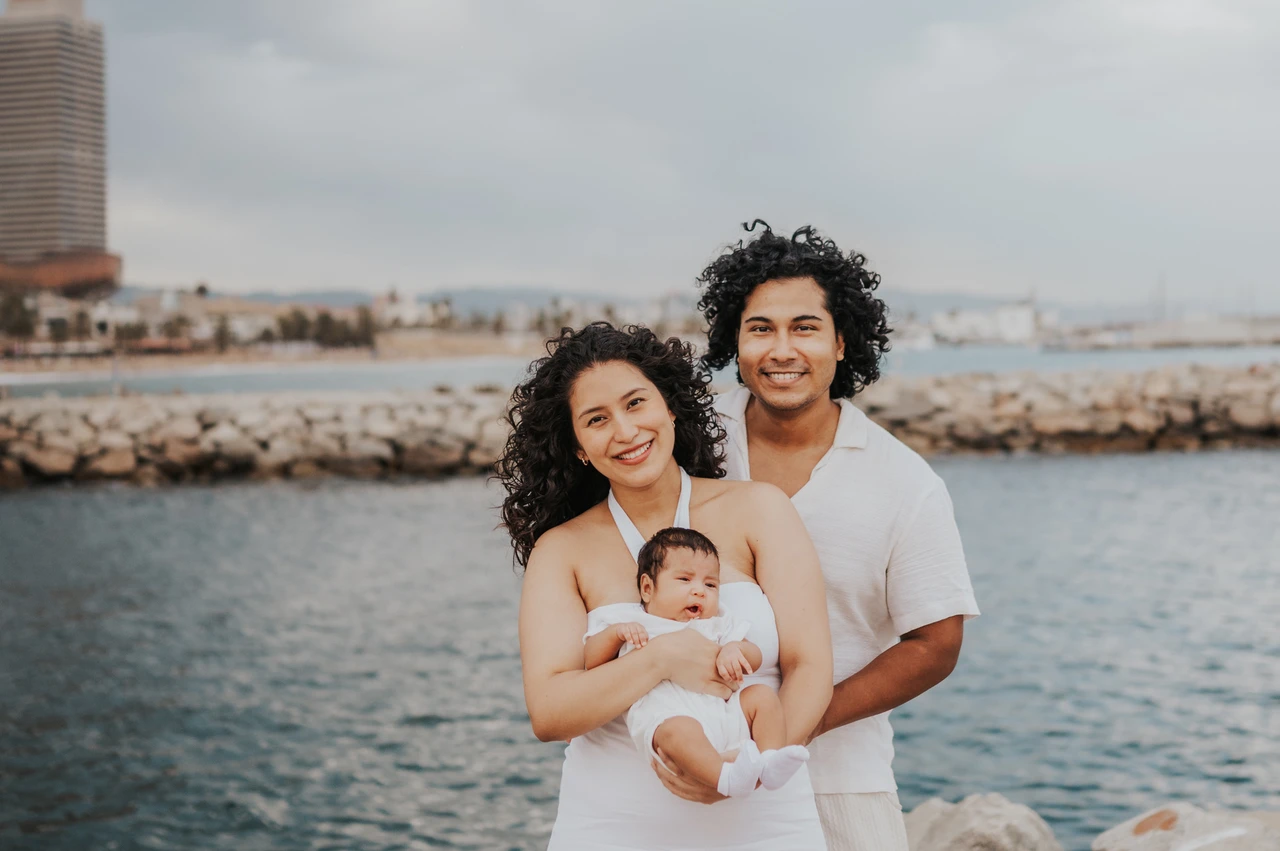 A family portrait with the city behind them | Barcelona newborn family photoshoot The mother holding the newborn with the father close behind and the city across the water during their newborn family photoshoot in Barcelona.