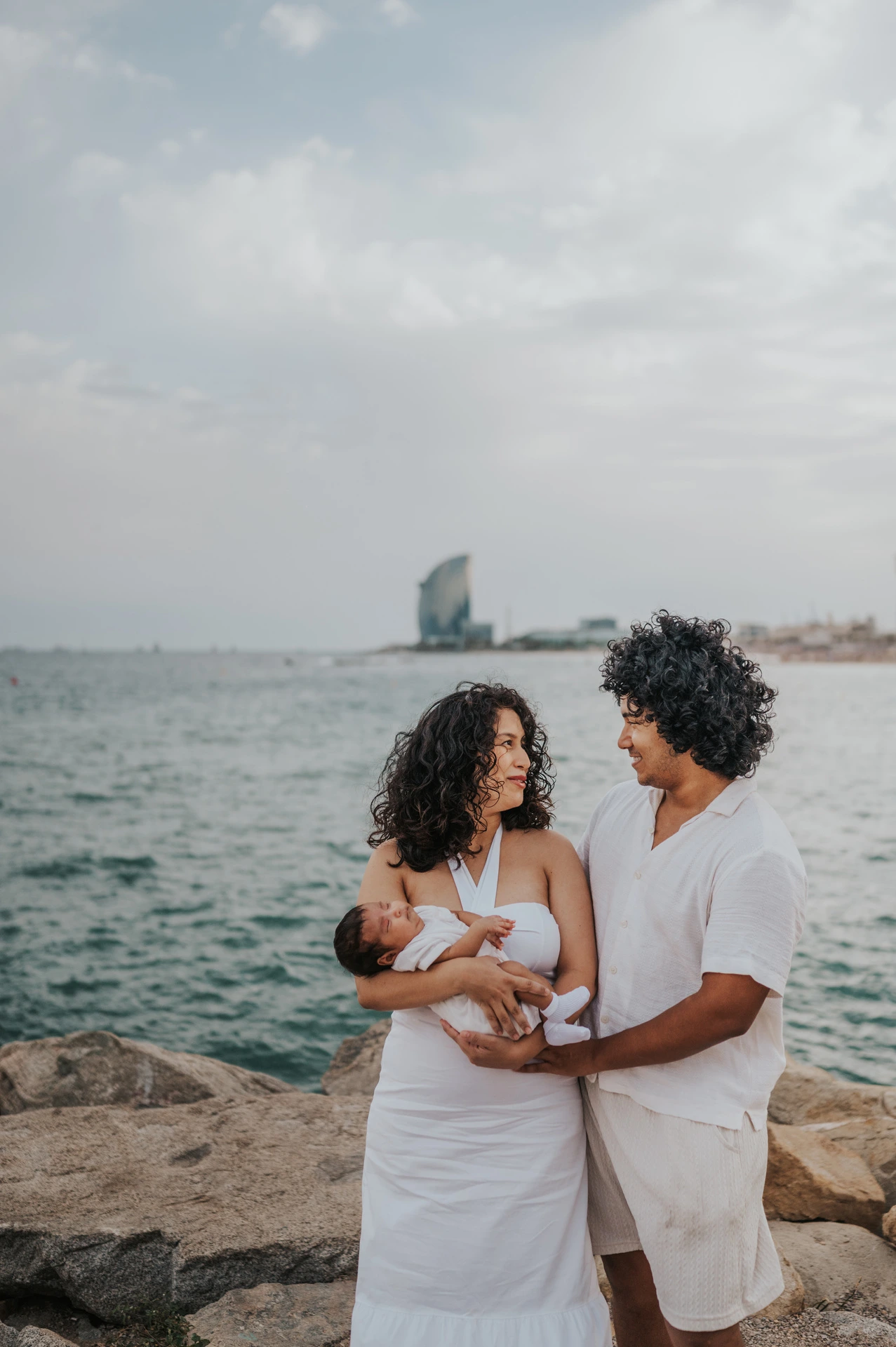 The parents looking towards each other while holding their newborn with the sea behind them during their newborn family photoshoot in Barcelona.