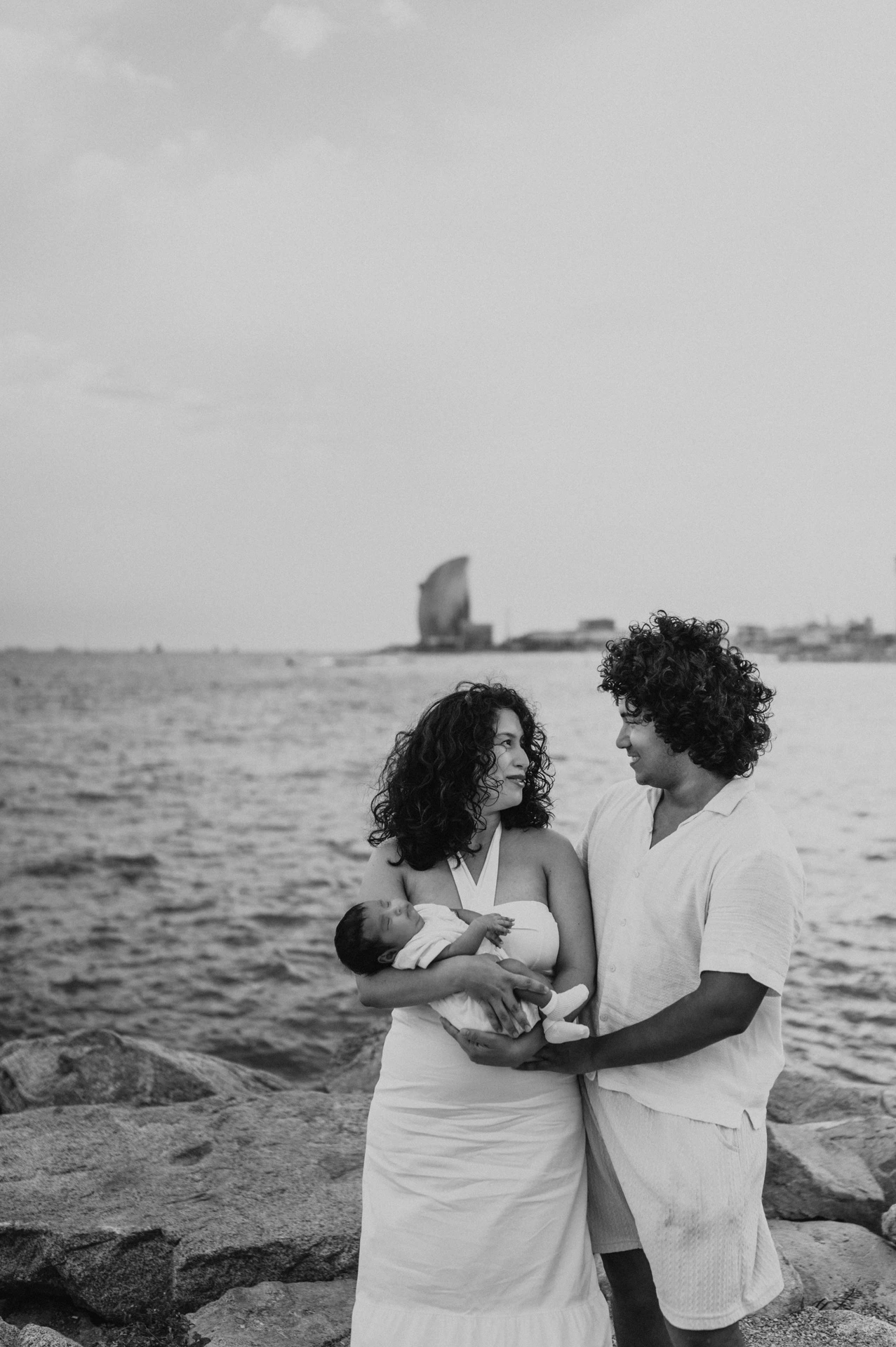 Black-and-white photograph: the parents looking towards each other while holding their newborn with the sea behind them during their newborn family photoshoot in Barcelona.