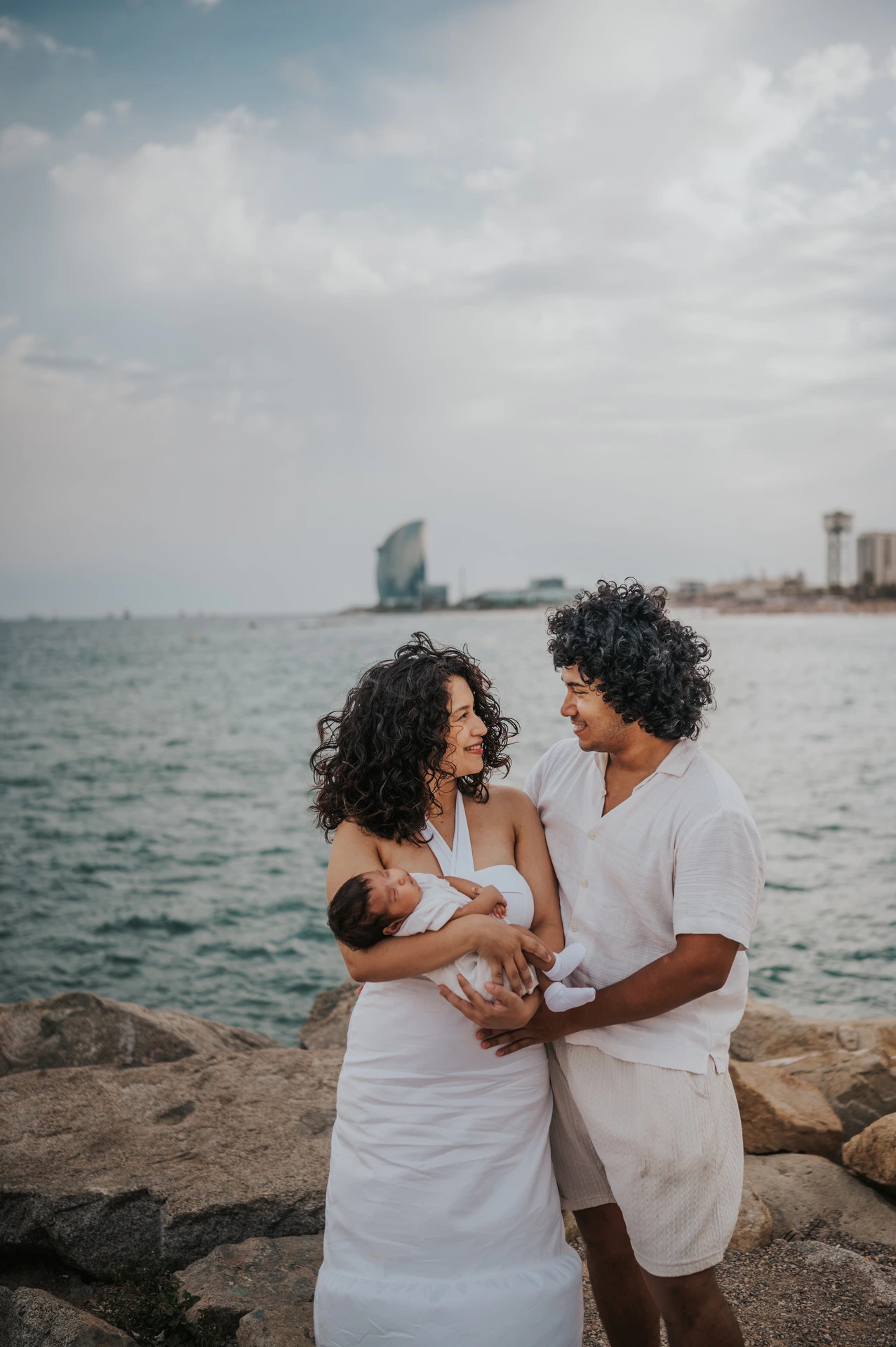 The parents looking towards each other while holding their newborn with the sea behind them during their newborn family photoshoot in Barcelona.
