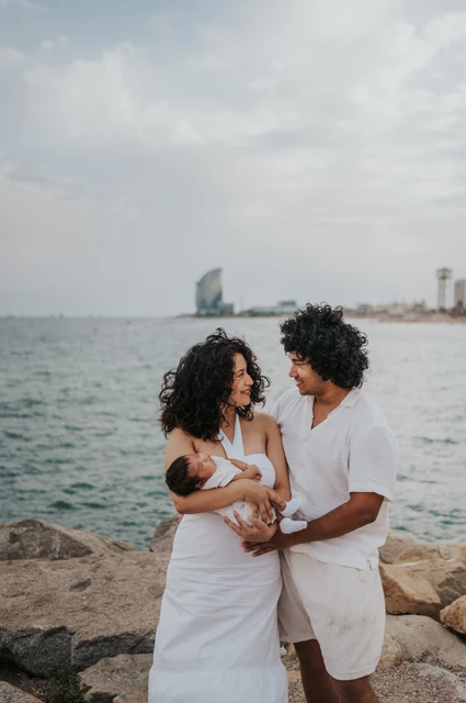 Together with their newborn by the sea | Barcelona newborn family photoshoot The parents looking towards each other while holding their newborn with the sea behind them during their newborn family photoshoot in Barcelona.
