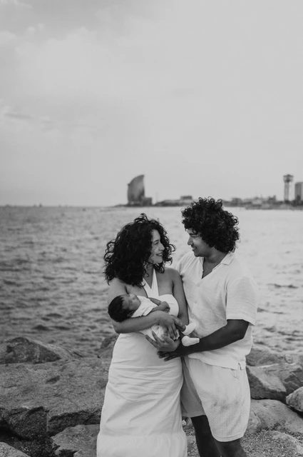 Together with their newborn by the sea in black and white | Barcelona newborn family photoshoot Black-and-white photograph: the parents looking towards each other while holding their newborn with the sea behind them during their newborn family photoshoot in Barcelona.