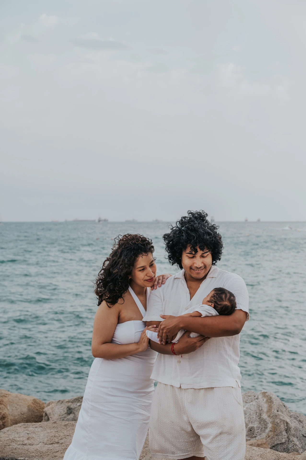 The father cradling the newborn while the mother stands close beside him by the water during their newborn family photoshoot in Barcelona.