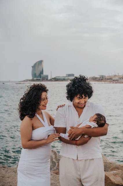 Their newborn in the father’s arms | Barcelona newborn family photoshoot The father cradling the newborn while the mother stands close beside him by the water during their newborn family photoshoot in Barcelona.