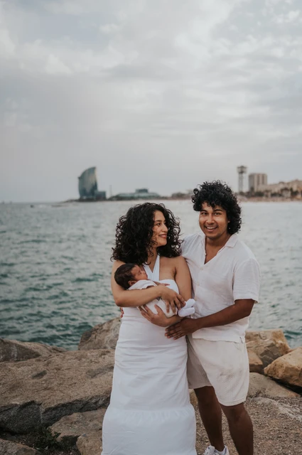 The father cradling the newborn while the mother stands close beside him by the water during their newborn family photoshoot in Barcelona.
