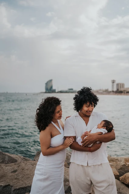 Their newborn in the father’s arms | Barcelona newborn family photoshoot The father cradling the newborn while the mother stands close beside him by the water during their newborn family photoshoot in Barcelona.