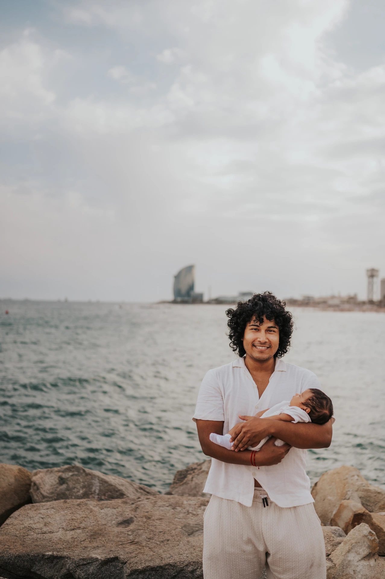 The father holding his newborn with the water and city behind him during their newborn family photoshoot in Barcelona.