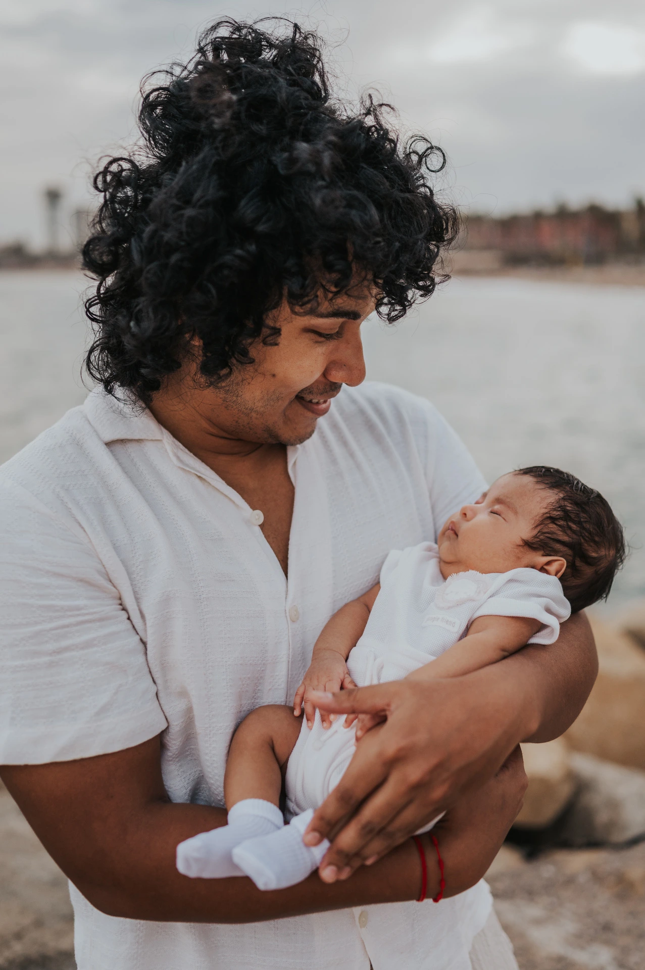 The father holding his newborn with the water and city behind him during their newborn family photoshoot in Barcelona.