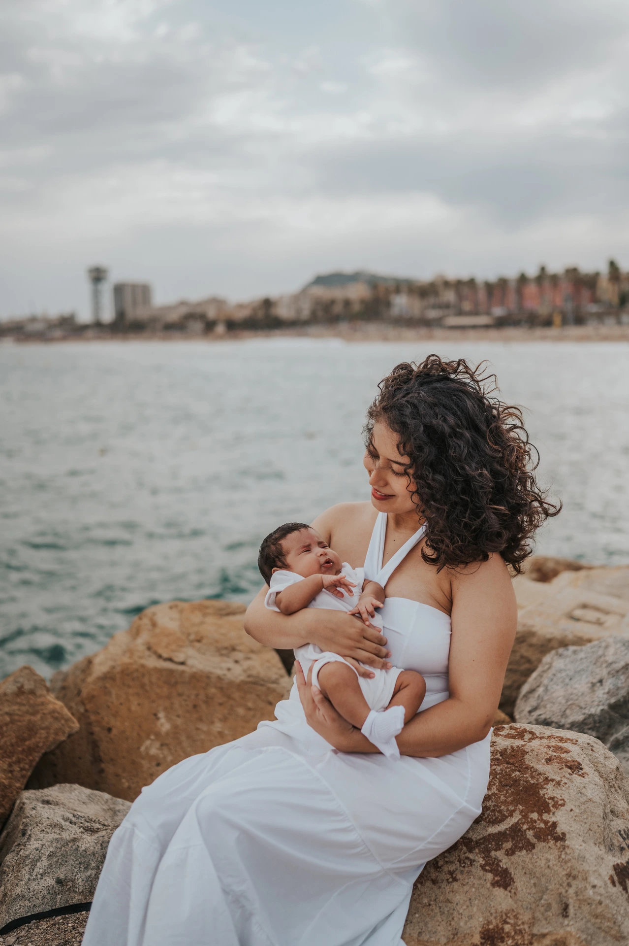 The mother sitting on a rock while holding her newborn beside the sea during their newborn family photoshoot in Barcelona.