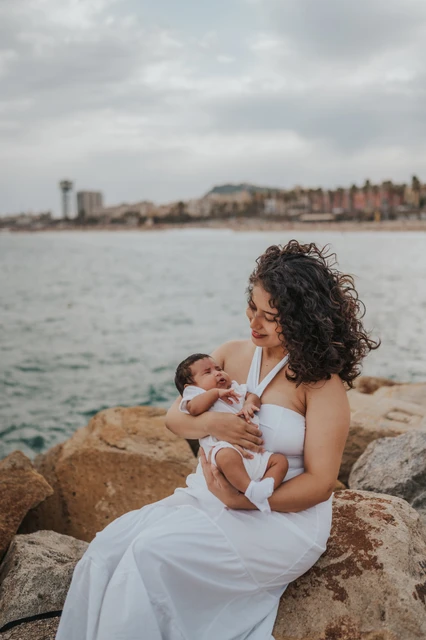 Mother and newborn seated by the shore | Barcelona newborn family photoshoot The mother sitting on a rock while holding her newborn beside the sea during their newborn family photoshoot in Barcelona.
