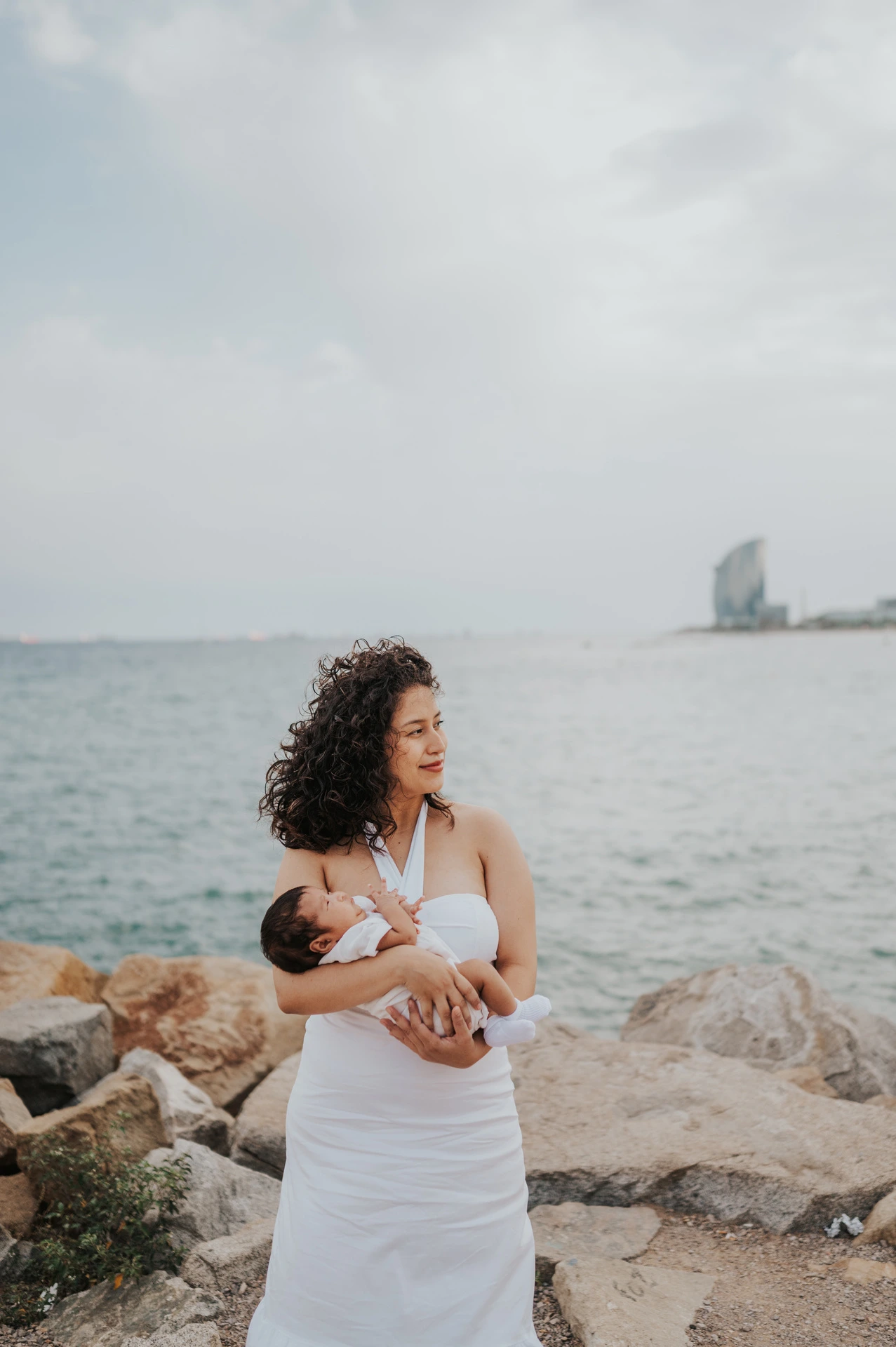 The mother cradling her newborn beside the rocky shoreline during their newborn family photoshoot in Barcelona.