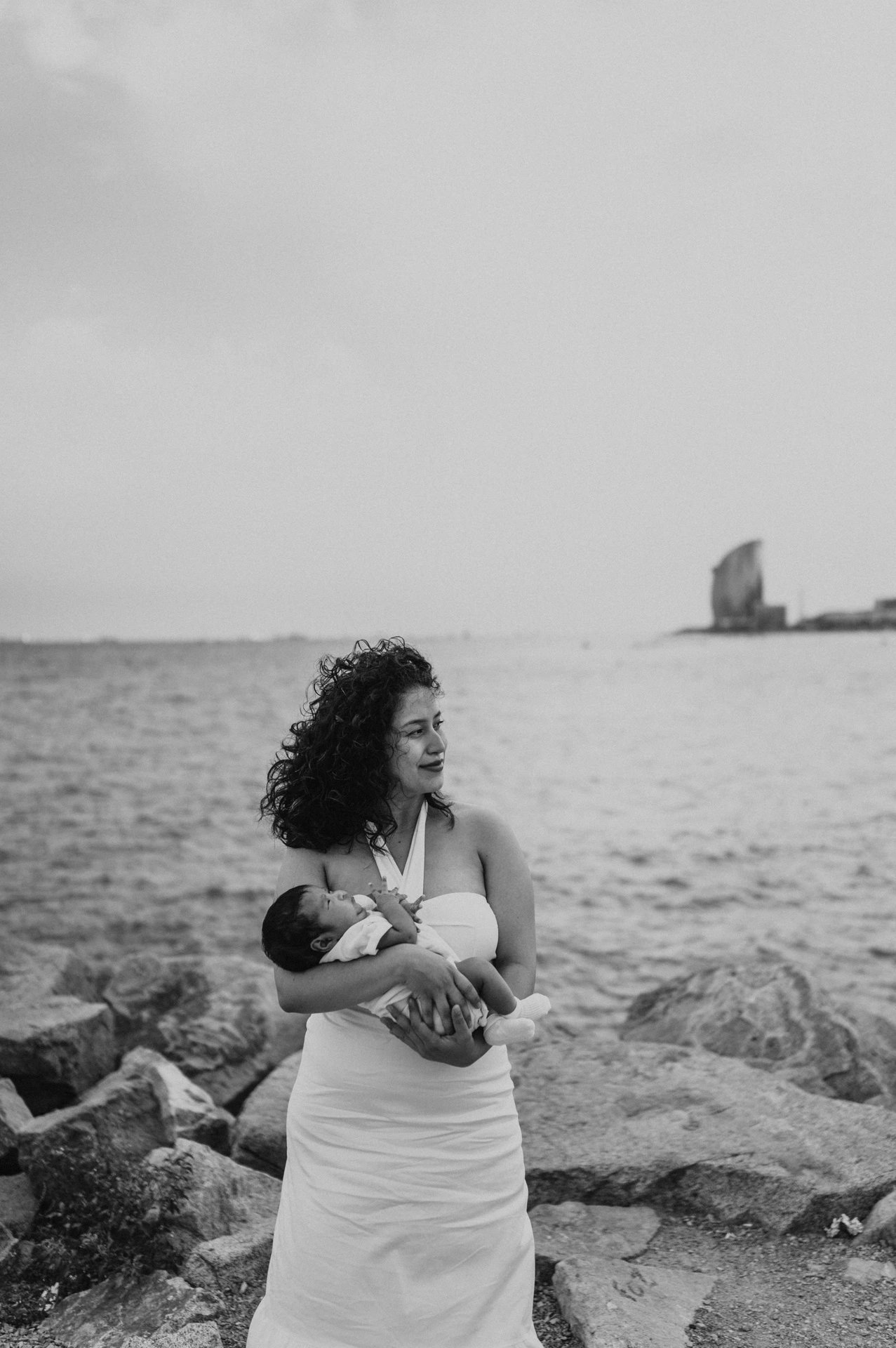 Black-and-white photograph: the mother cradling her newborn beside the rocky shoreline during their newborn family photoshoot in Barcelona.