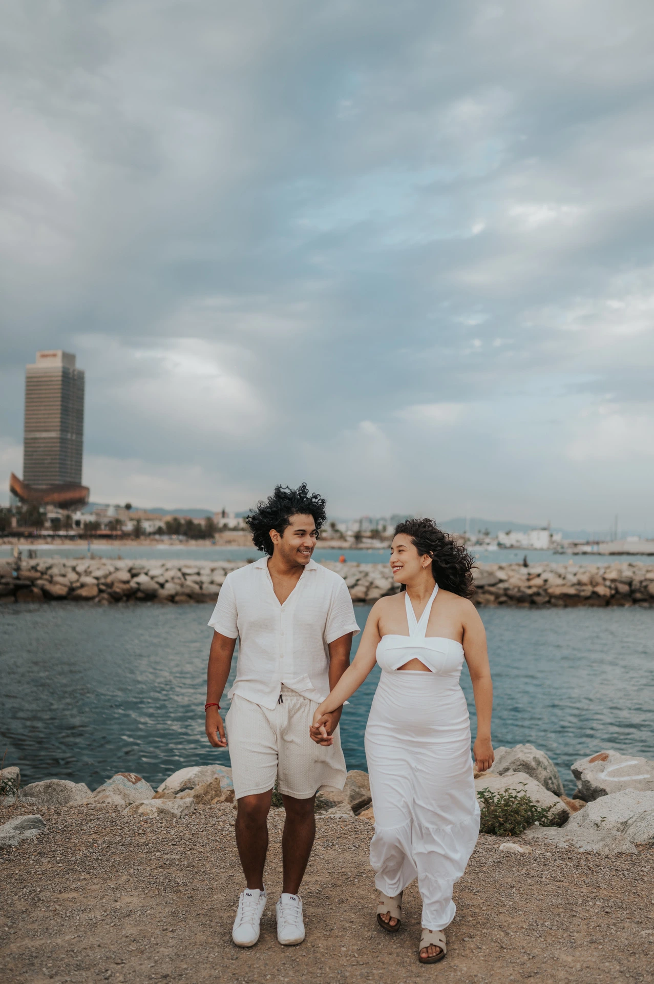 The parents walking hand in hand along the shore with the city behind them during their newborn family photoshoot in Barcelona.