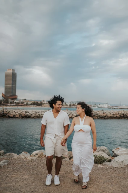 The parents walking hand in hand along the shore with the city behind them during their newborn family photoshoot in Barcelona.