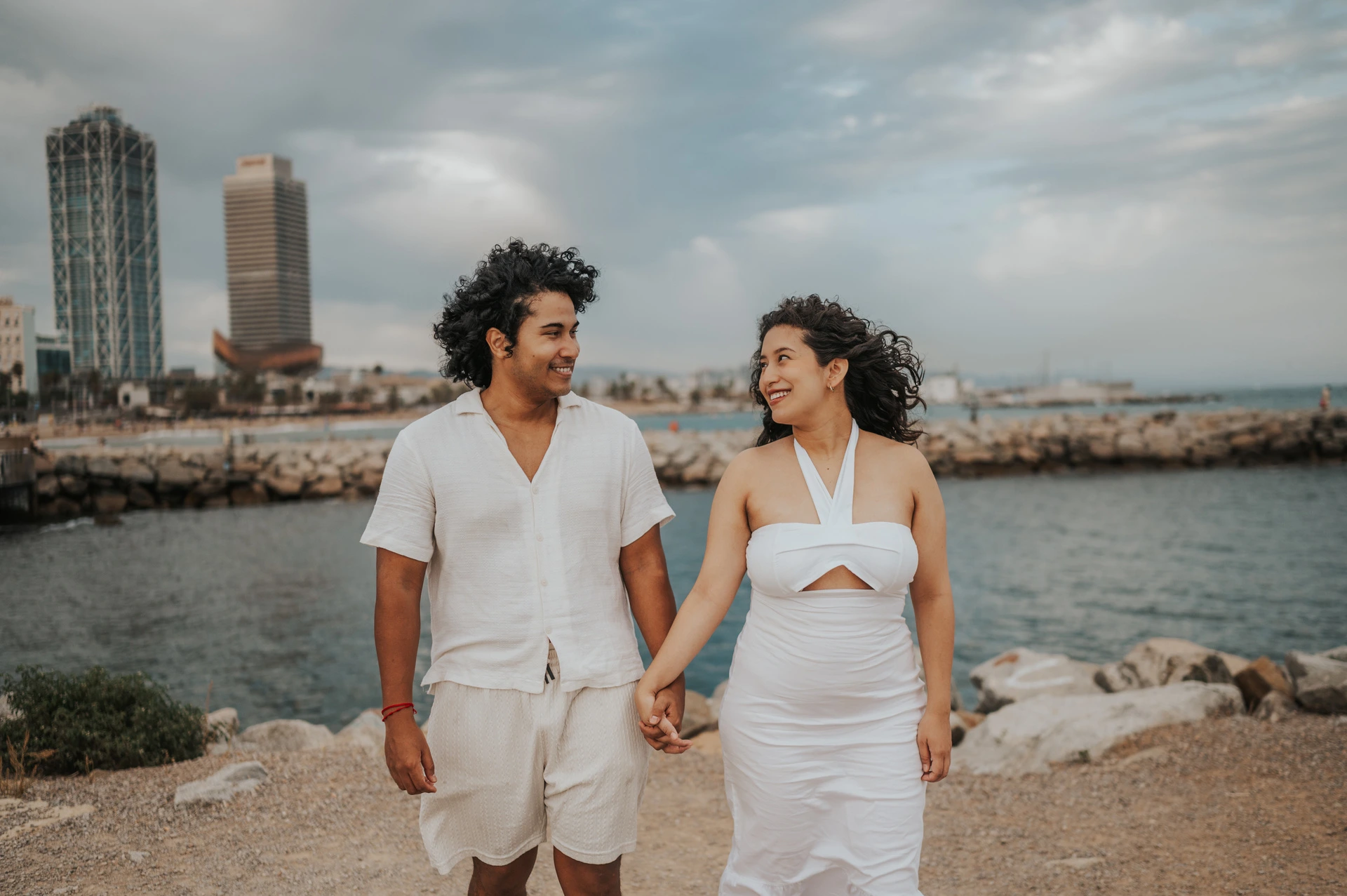 The parents walking hand in hand along the shore with the city behind them during their newborn family photoshoot in Barcelona.