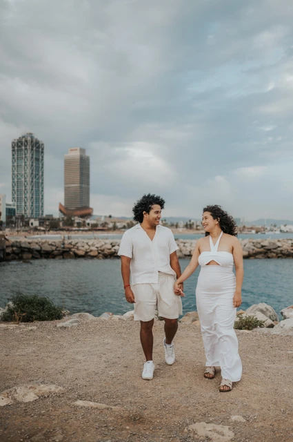 A walk together beside the shoreline | Barcelona newborn family photoshoot The parents walking hand in hand along the shore with the city behind them during their newborn family photoshoot in Barcelona.