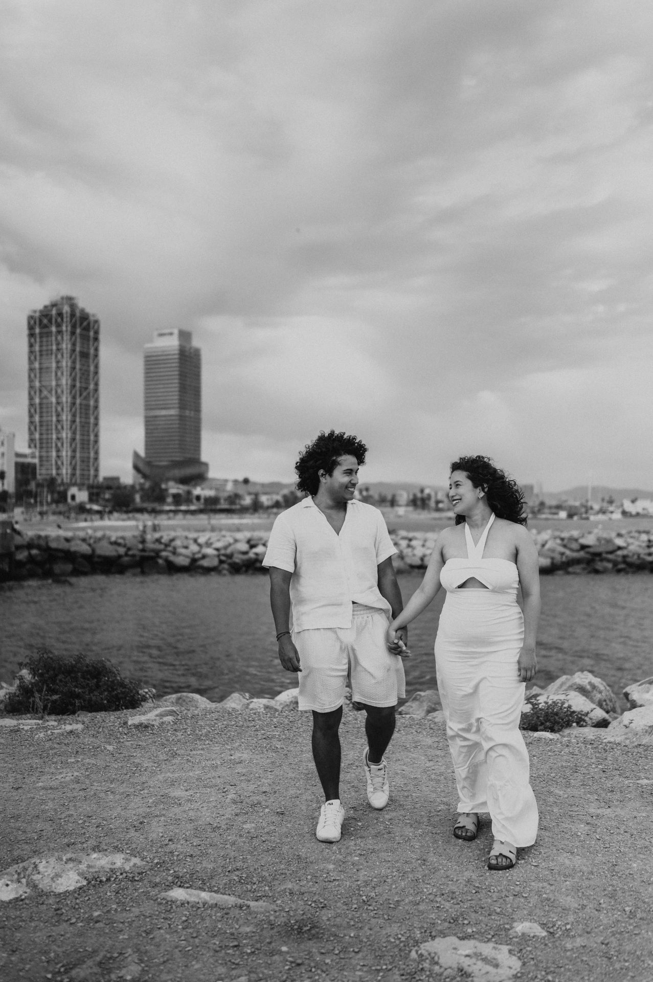 Black-and-white photograph: the parents walking hand in hand along the shore with the city behind them during their newborn family photoshoot in Barcelona.