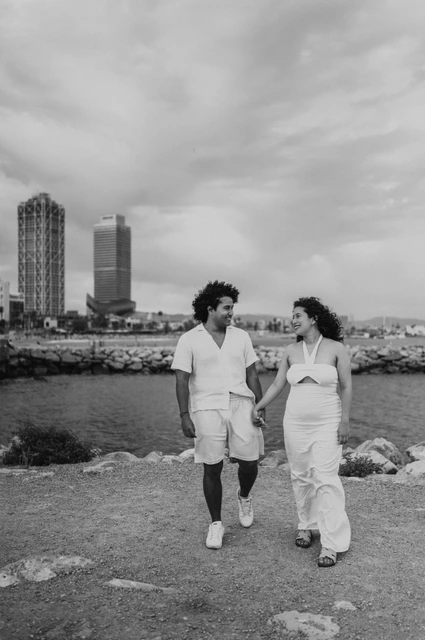 A walk together beside the shoreline in black and white | Barcelona newborn family photoshoot Black-and-white photograph: the parents walking hand in hand along the shore with the city behind them during their newborn family photoshoot in Barcelona.