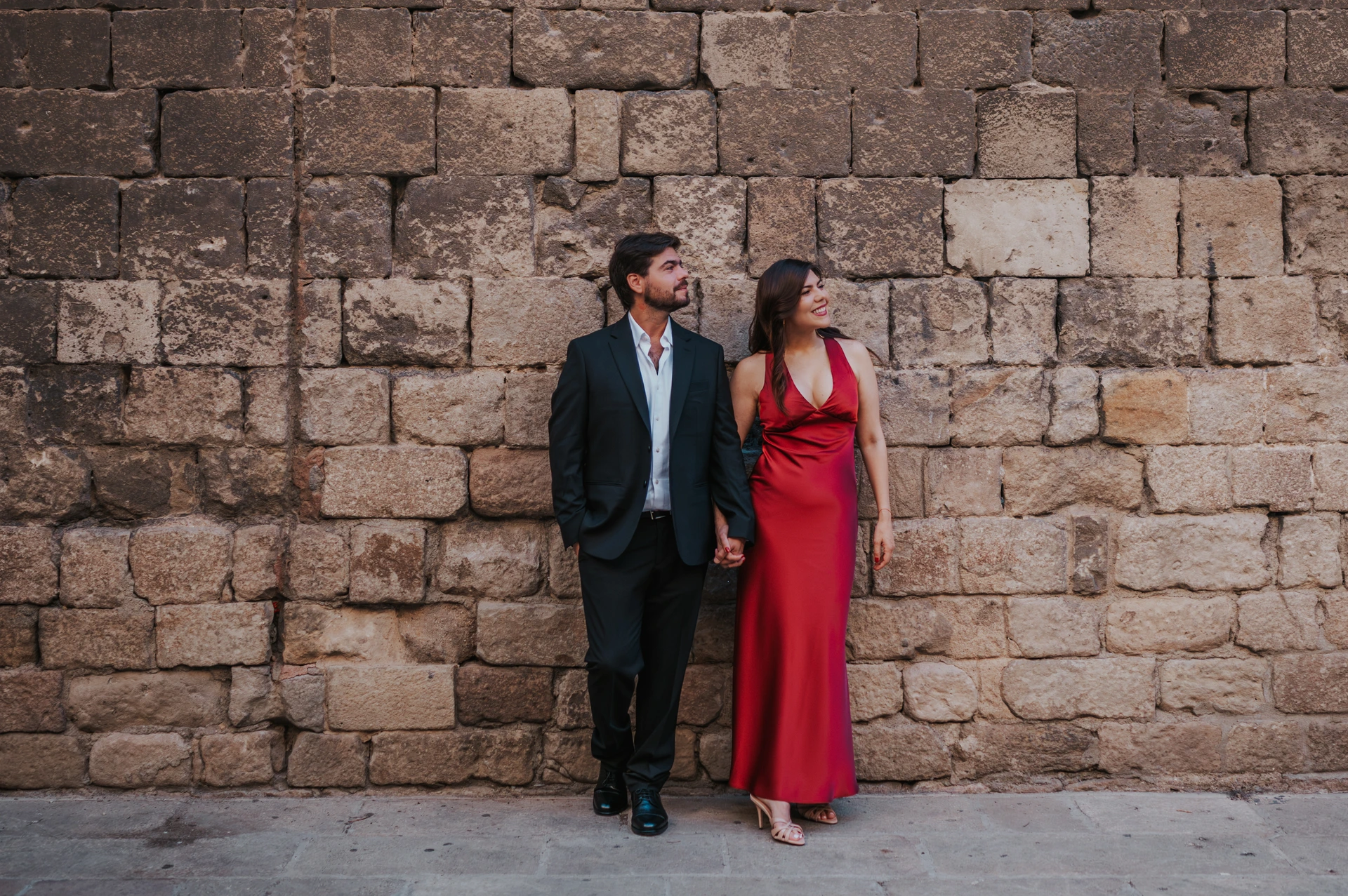 Two adults stand together against an old stone wall during a Barcelona photoshoot, with their full outfits visible.