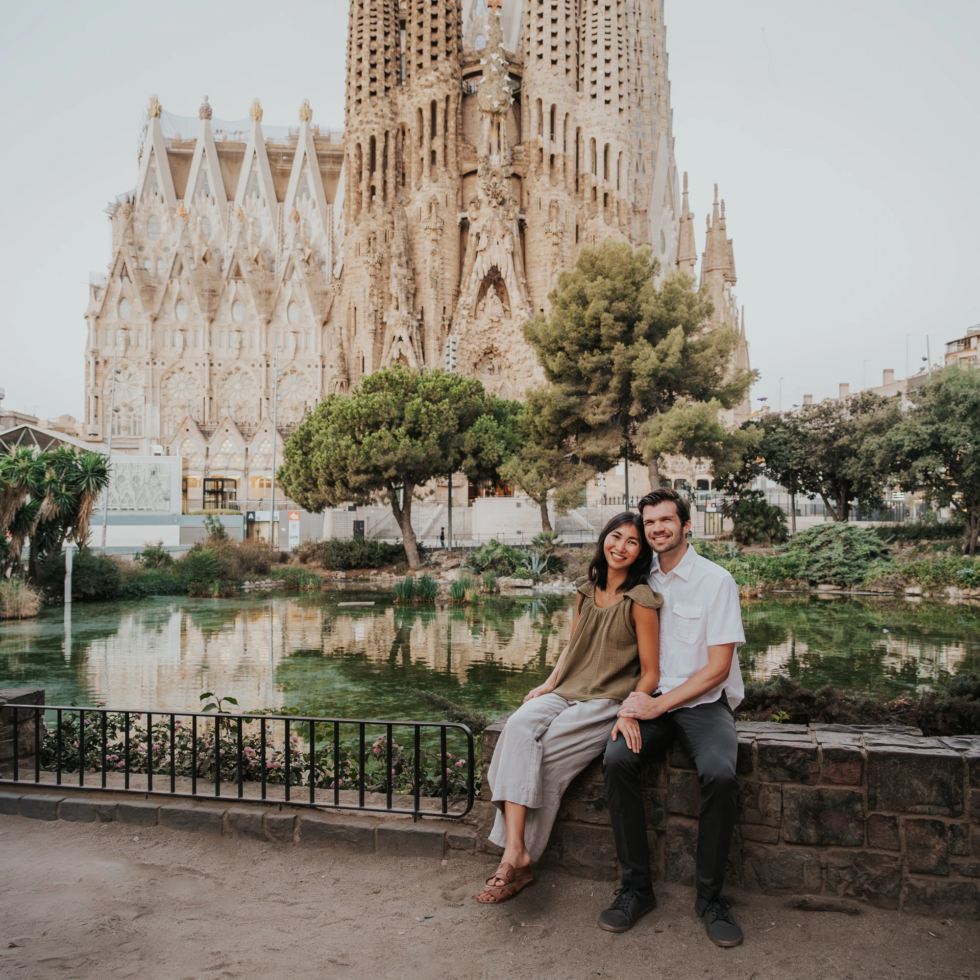 Barcelona Photoshoot Duration - Sagrada Familia Portrait Two adults sit beside the Sagrada Familia pond during a focused Barcelona photoshoot, with the basilica rising behind them.
