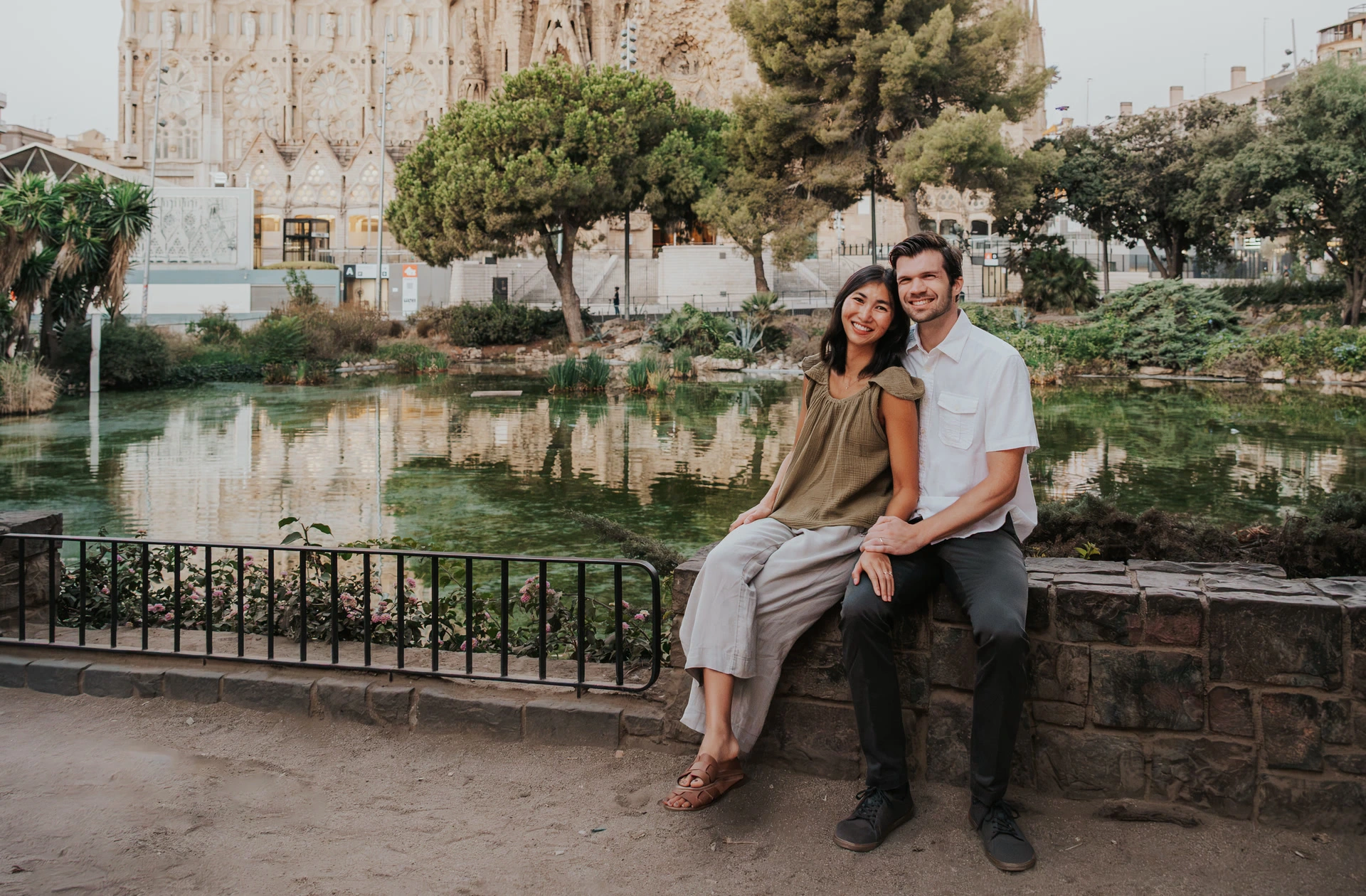 Two adults sit beside the pond at Sagrada Família, with the basilica's sculpted exterior rising behind their Barcelona portrait.
