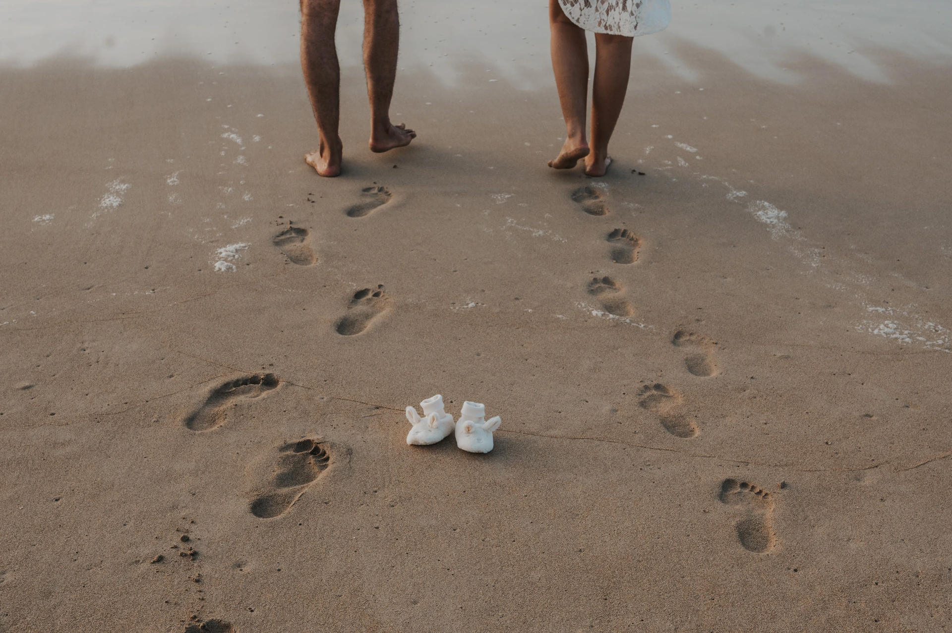 A close-up of baby shoes and footprints on Dubai sand during a maternity shoot captures styling context and storytelling details around the parents’ outfits.