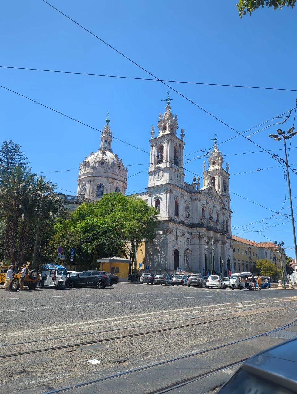 Lisbon Tram Photoshoot - Estrela Basilica Estrela Basilica's pale dome and ornate towers rise behind trees, tram rails, overhead wires and a broad Lisbon street.