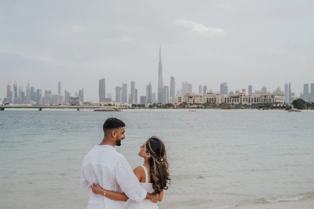 A couple embraces on a Dubai shoreline with calm water and skyline views behind them during a beach proposal session.