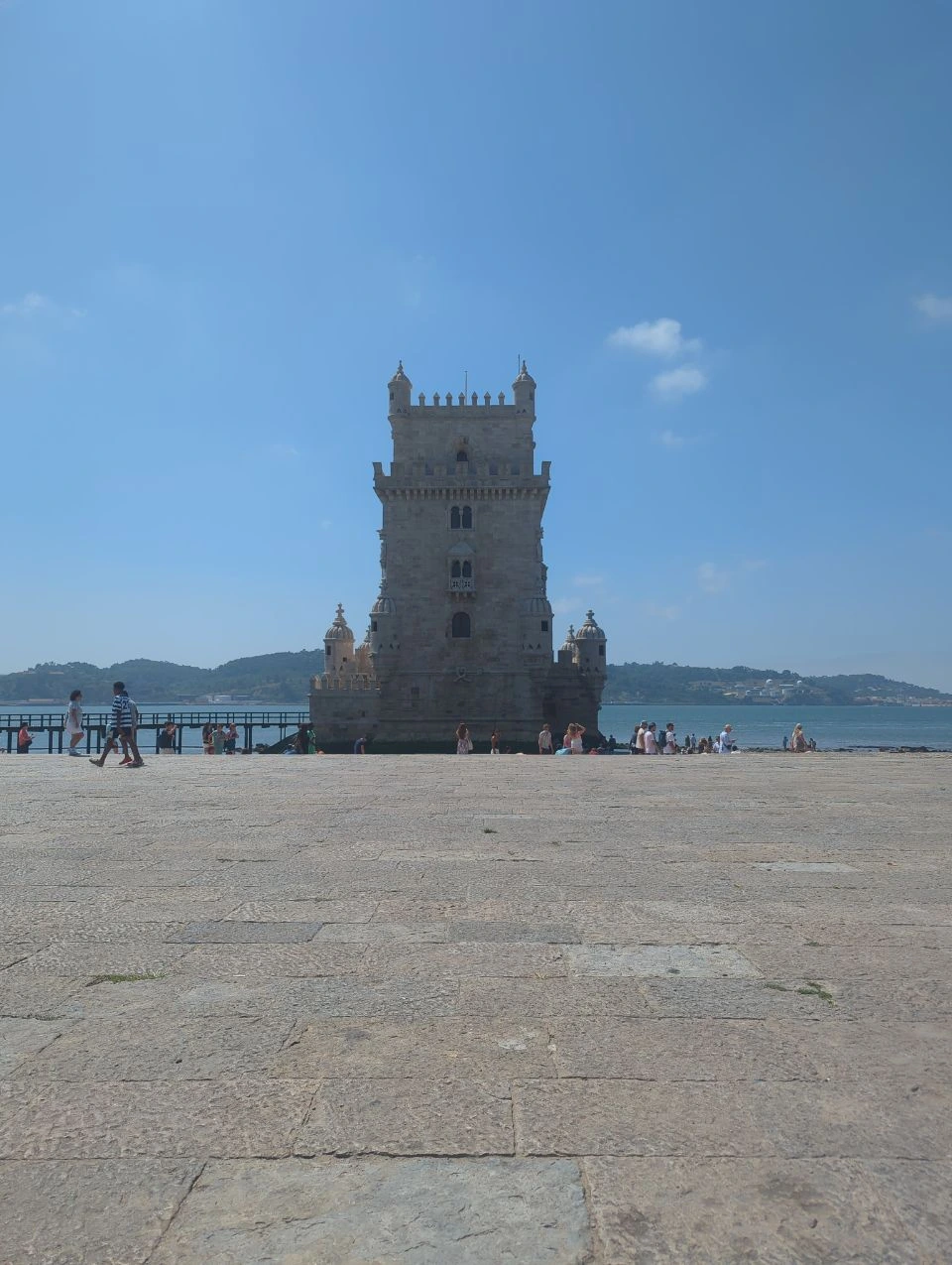 Belém Tower rises beyond a broad stone plaza, with small groups of visitors beside the Tagus waterfront.