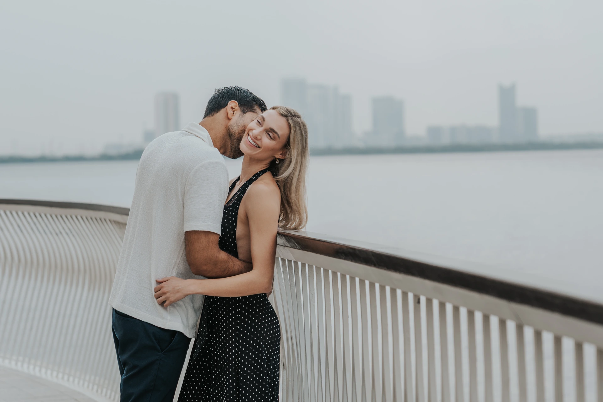 A couple hugs along a Dubai waterfront railing with city skyline towers softly visible in the background.