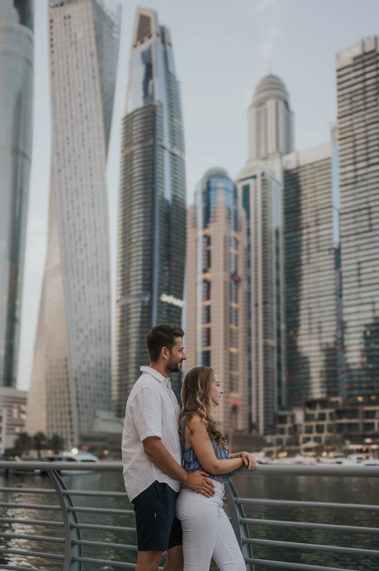 A couple stands in a modern downtown Dubai setting with high-rise buildings and curved architecture behind them.