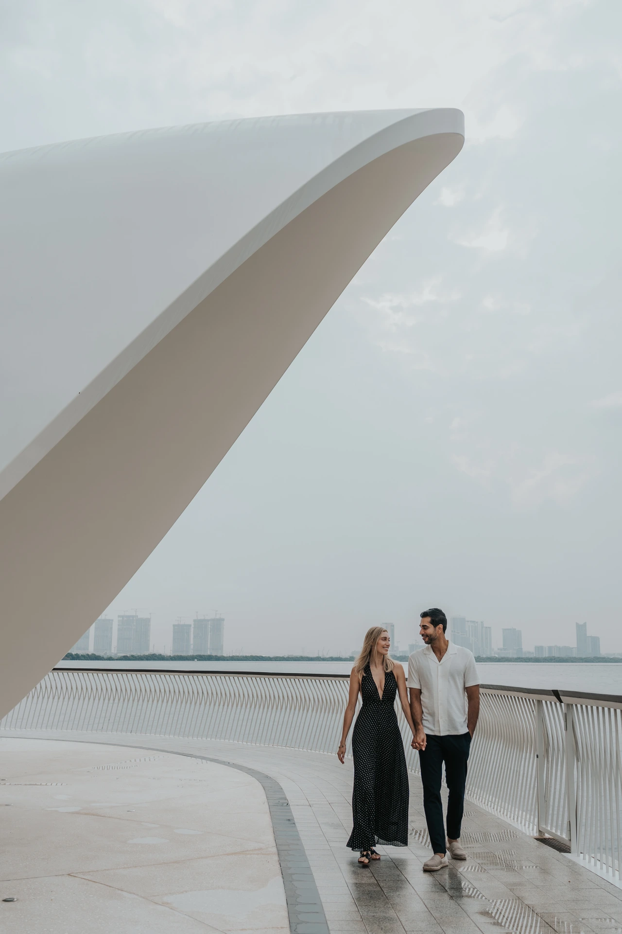 A couple stands beneath a clean modern architectural frame near the waterfront with Dubai skyline in the distance.