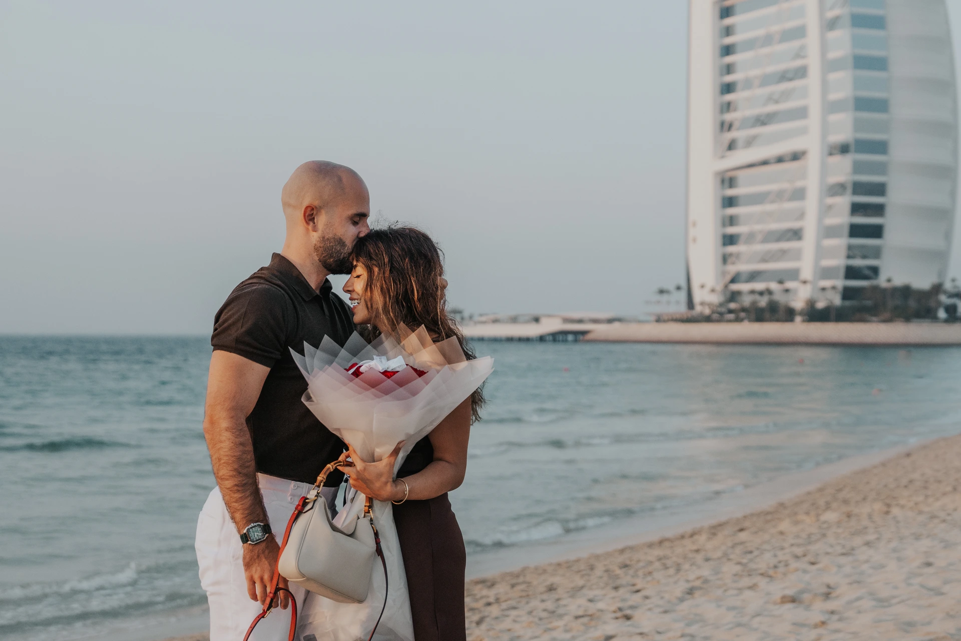 A couple embraces on Dubai beach after a Shimmers proposal while holding a bouquet, with Burj Al Arab visible in the background.