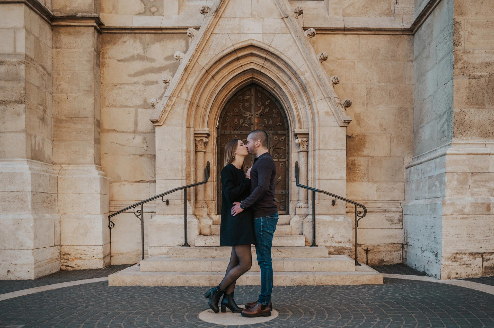 A landscape portrait of the couple embracing beneath a pointed church doorway near Fisherman’s Bastion during their couple photoshoot in Budapest.
