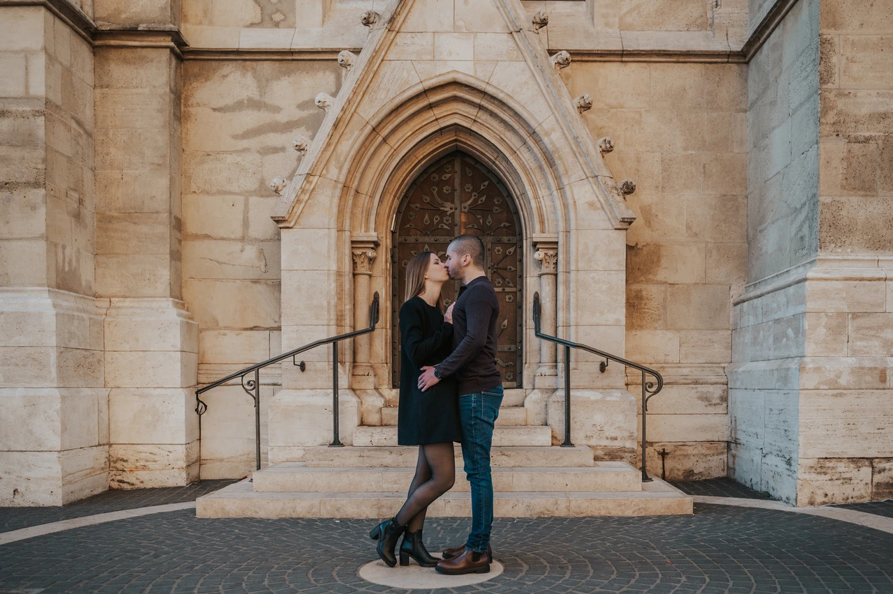 A romantic portrait beneath the pointed doorway at a Budapest couple photoshoot A landscape portrait of the couple embracing beneath a pointed church doorway near Fisherman’s Bastion during their couple photoshoot in Budapest.