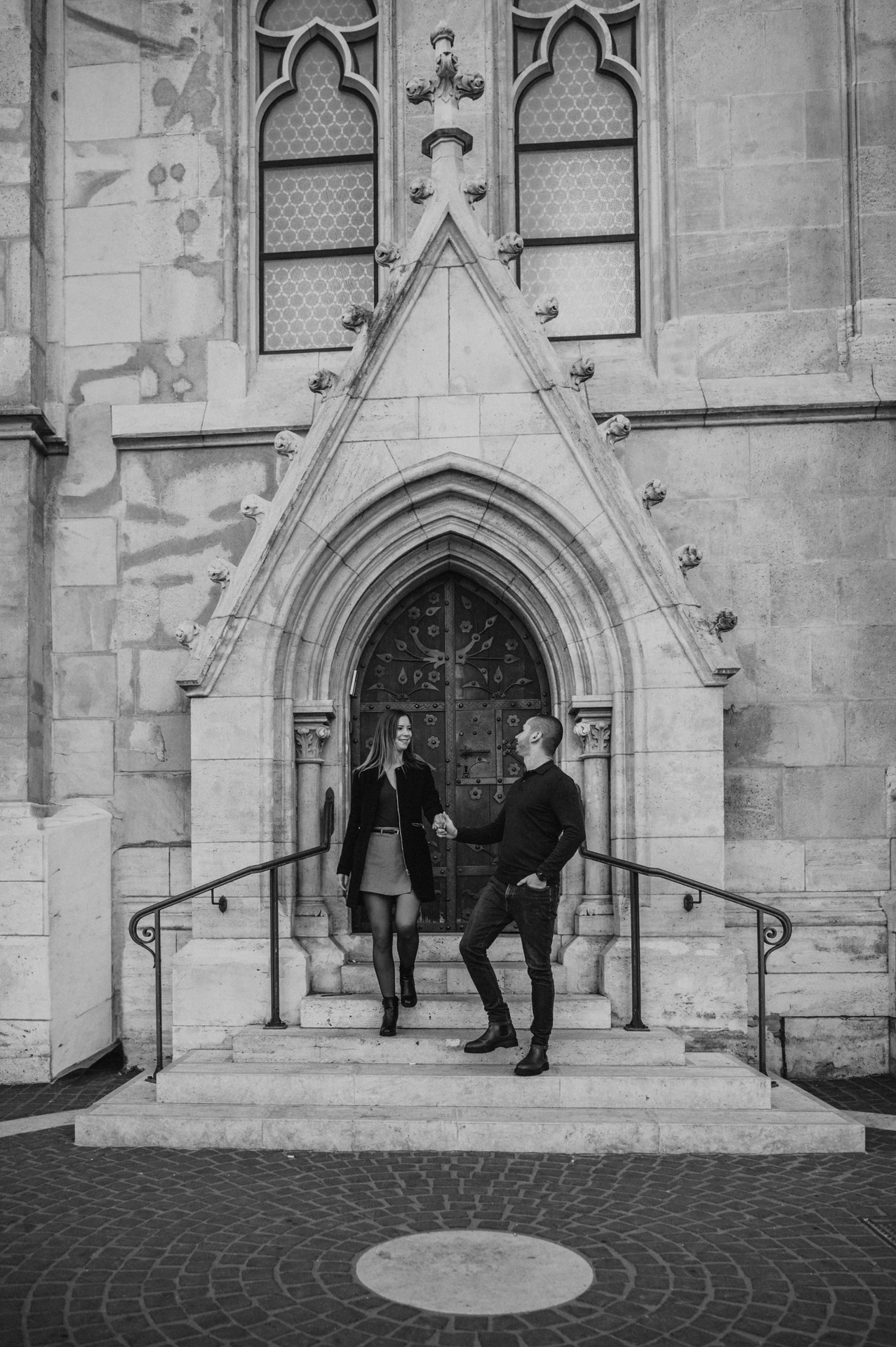 Black-and-white portrait of the couple walking hand in hand down the steps of a pointed church doorway near Fisherman’s Bastion during their couple photoshoot in Budapest.