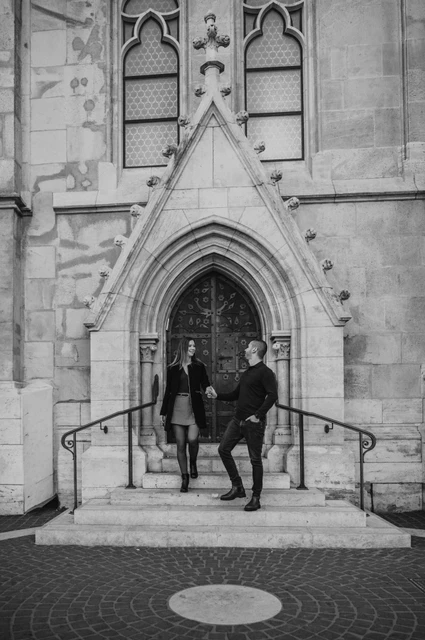 A doorway walk in black and white at a Budapest couple photoshoot Black-and-white portrait of the couple walking hand in hand down the steps of a pointed church doorway near Fisherman’s Bastion during their couple photoshoot in Budapest.