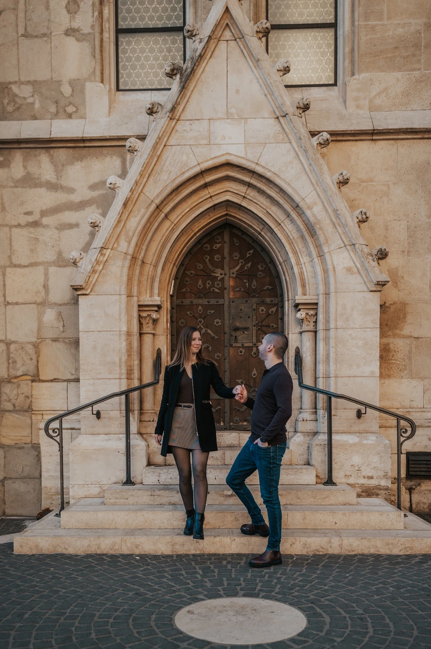 A walk beside the carved church doorway at a Budapest couple photoshoot The couple walking hand in hand down the steps beneath a carved church doorway near Fisherman’s Bastion during their couple photoshoot in Budapest.