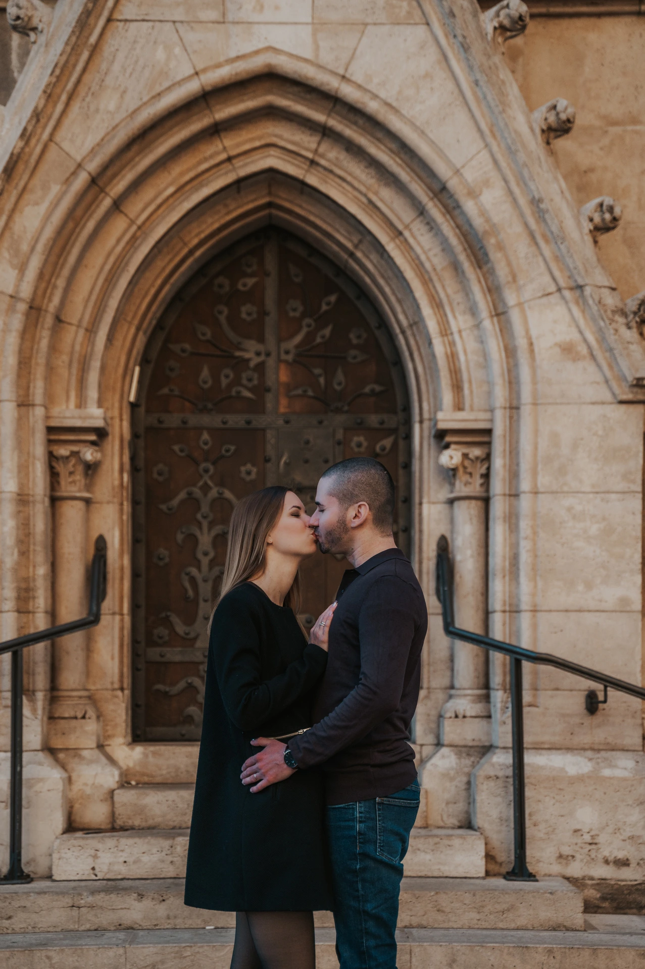 A close portrait of the couple embracing in front of an ornate wooden door beneath a pointed stone arch near Fisherman’s Bastion during their couple photoshoot in Budapest.
