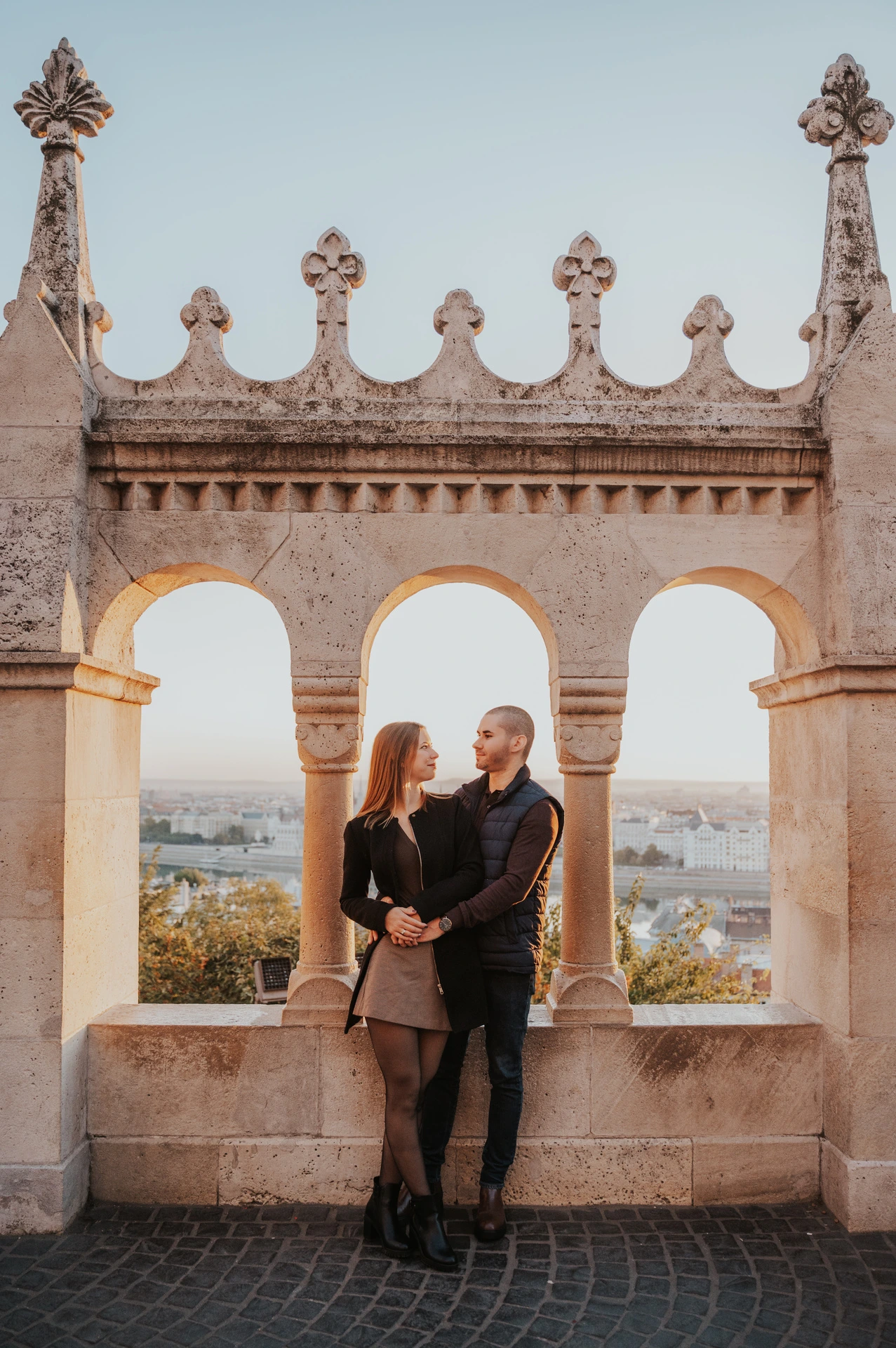The couple embracing beneath the carved stone arches at Fisherman’s Bastion with Parliament beyond them during their couple photoshoot in Budapest.