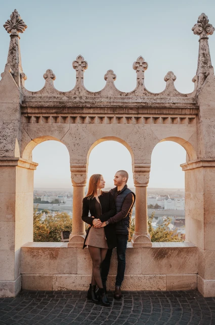 An embrace beneath the carved arches at a Budapest couple photoshoot The couple embracing beneath the carved stone arches at Fisherman’s Bastion with Parliament beyond them during their couple photoshoot in Budapest.