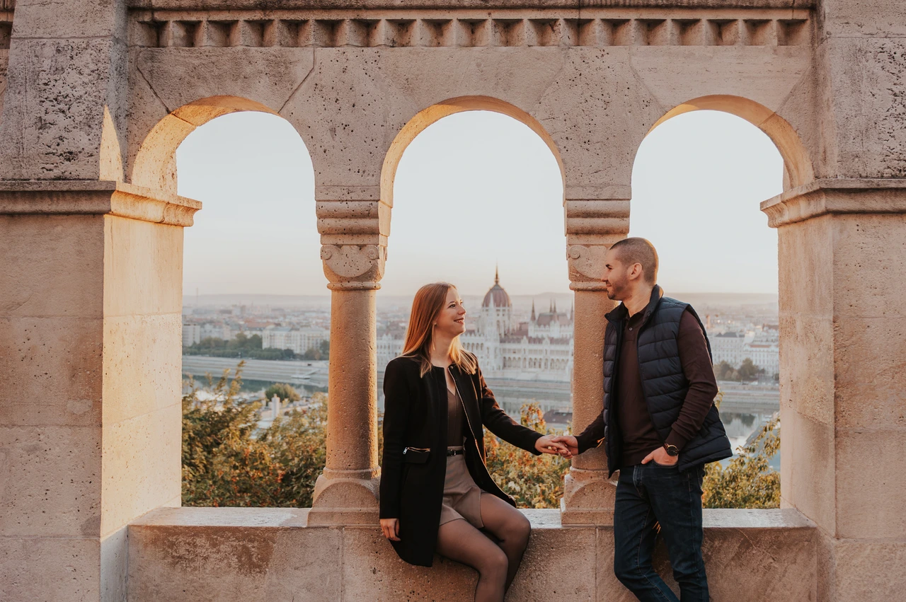 Holding hands beneath the Bastion arches at a Budapest couple photoshoot A landscape portrait of the couple holding hands beneath the stone arches at Fisherman’s Bastion with Parliament in the background during their couple photoshoot in Budapest.