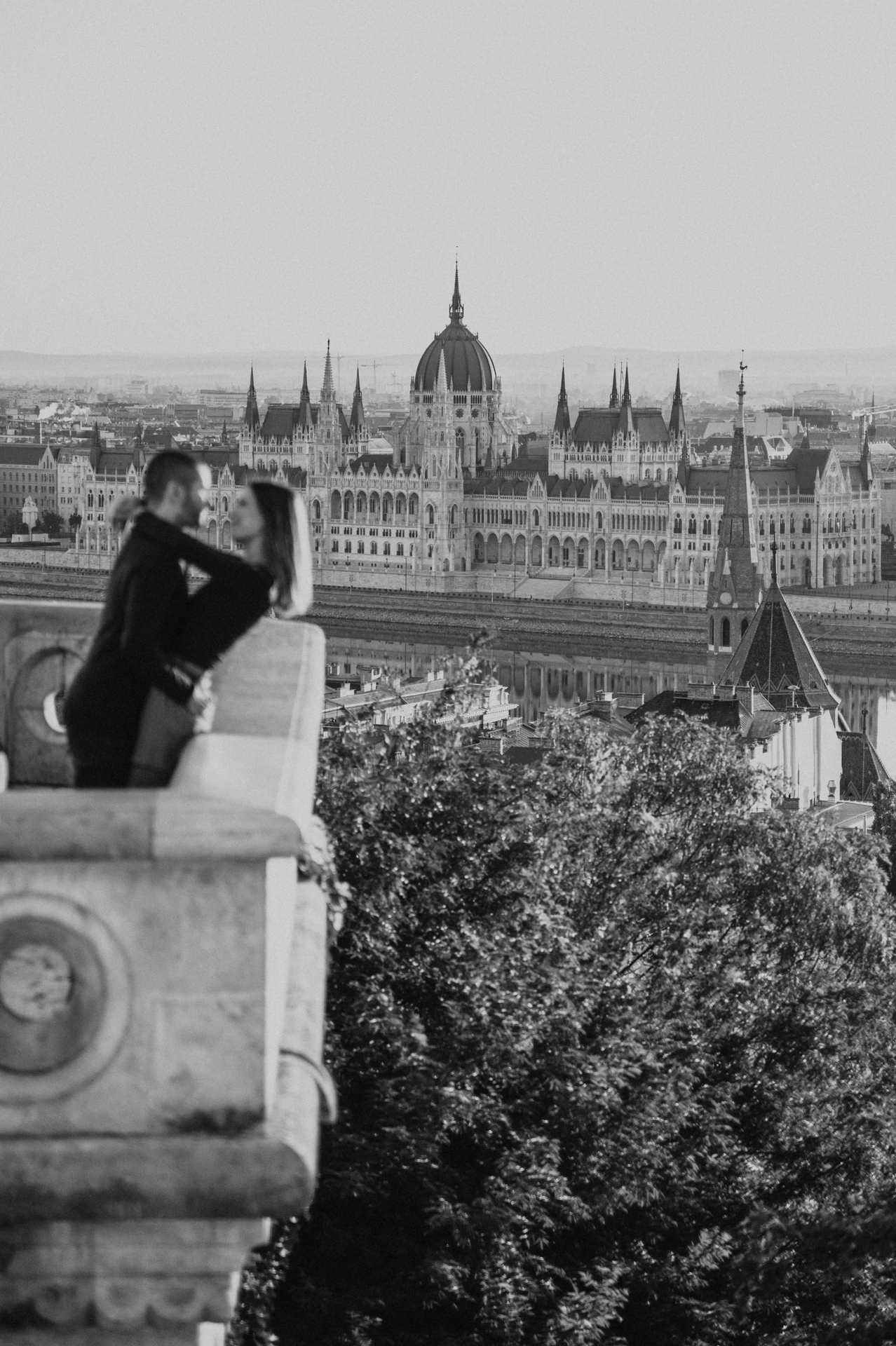 Black-and-white close portrait of the couple embracing beside Fisherman’s Bastion’s balcony with Parliament behind them during their couple photoshoot in Budapest.