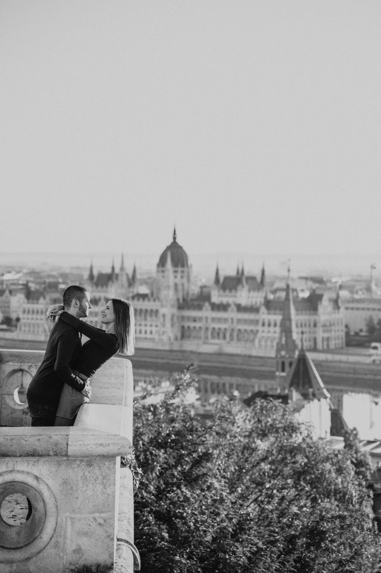 Black-and-white portrait of the couple embracing on Fisherman’s Bastion’s balcony with Parliament behind them during their couple photoshoot in Budapest.