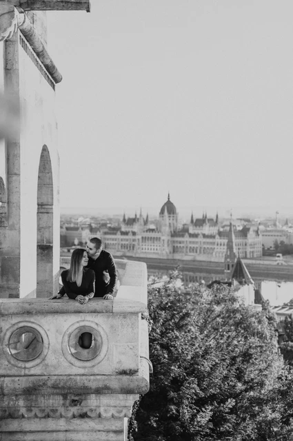 A romantic balcony portrait in black and white at a Budapest couple photoshoot Black-and-white portrait of the couple leaning close over Fisherman’s Bastion’s carved stone balcony with Parliament in the background during their couple photoshoot in Budapest.