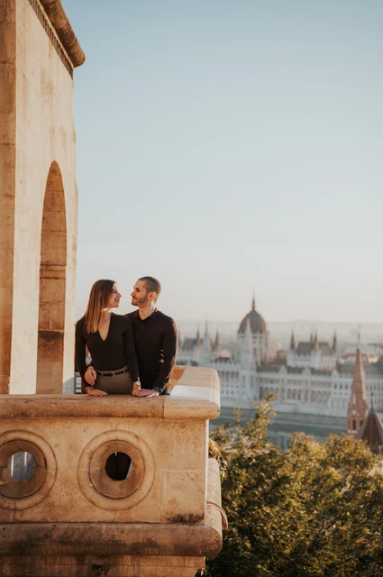 A quiet moment with Parliament behind them at a Budapest couple photoshoot The couple looking at one another behind a carved stone balcony at Fisherman’s Bastion with Parliament across the Danube during their couple photoshoot in Budapest.