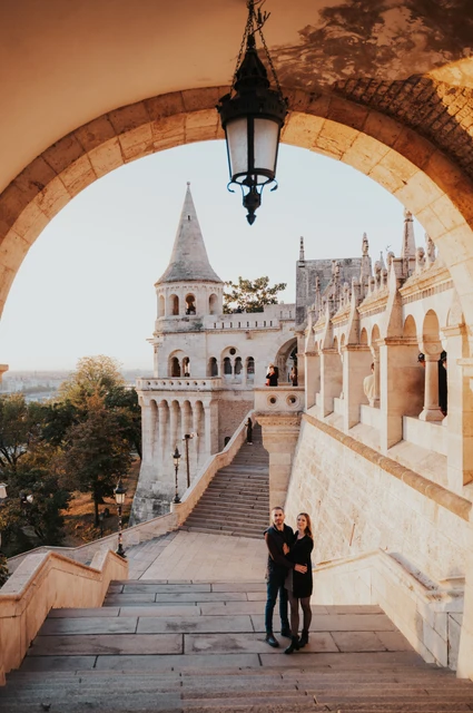 A grand arch above the Bastion stairs at a Budapest couple photoshoot A wide portrait of the couple beneath a large stone arch and hanging lamp on the stairs at Fisherman’s Bastion during their couple photoshoot in Budapest.